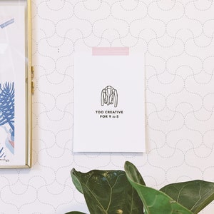 Op de afbeelding: Een wit papier met een zwarte lijntekening van een jas en de tekst "TOO CREATIVE FOR 9 to 5" is op een behang geplakt. Een ingelijste foto en een groene plant zijn ook zichtbaar.