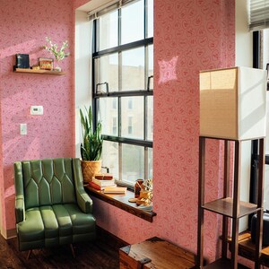 May include: A pink room with a green armchair, a wooden chest, and a floor lamp. The walls are covered in a pink wallpaper with a repeating pattern of hearts with the word "Baby" inside each heart.