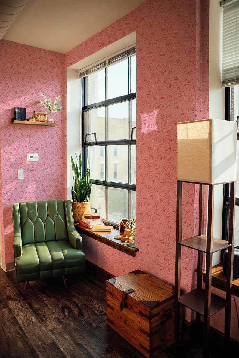 May include: A pink room with a green armchair, a wooden chest, and a floor lamp. The walls are covered in a pink wallpaper with a repeating pattern of hearts with the word "Baby" inside each heart.