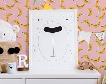 May include: A white framed print with a black and white illustration of a bear face on a pink background with yellow banana shapes. A white stuffed animal, a white ceramic cup with a black bear face, and a white wooden letter R are on a white table in front of the print.