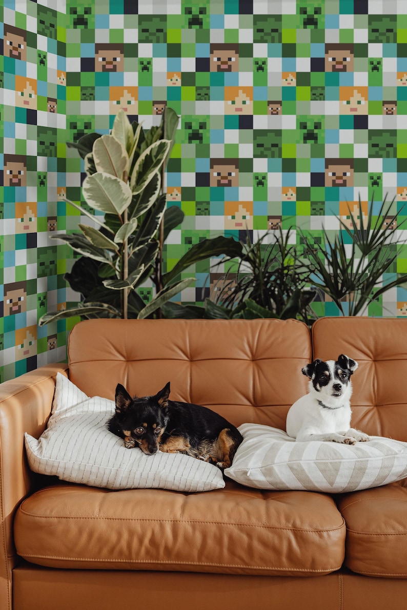 May include: Two dogs, one black and one white, are sitting on a brown leather couch in front of a wall covered in a green, blue, and white Minecraft-themed wallpaper.