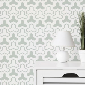 Op de afbeelding: Een wit nachtkastje met een lade en een lamp erop. De lamp heeft een witte kap en een witte voet. Een plant in een pot met groene bladeren staat rechts van het nachtkastje. De muur achter het nachtkastje heeft een geometrisch patroon van grijze driehoeken.