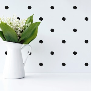 May include: A white pitcher filled with a bouquet of white lily of the valley flowers. The pitcher is on a white surface in front of a white wall with a black polka dot pattern.