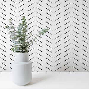 May include: A white ceramic vase with a rounded shape holds green leafy branches. The vase sits on a white surface in front of a white wall with a black chevron pattern. The image has a minimalist, modern aesthetic.