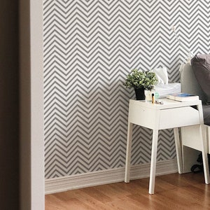 May include: A white nightstand with a drawer sits next to a bed with gray bedding and a white headboard. The wall behind the bed is covered in gray and white chevron patterned wallpaper.