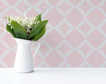May include: A white ceramic jug filled with a bouquet of white lily of the valley flowers and green leaves. The jug has a curved handle and a flared rim, standing against a light pink wall with a white geometric diamond pattern.