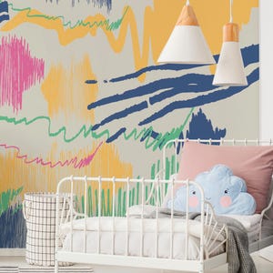 Puede incluir: Una cama de metal blanco con una almohada rosa y una almohada azul en forma de nube. La pared detrás de la cama tiene un diseño abstracto colorido con garabatos rosas, amarillos, azules y verdes. Una alfombra a rayas blancas y negras está frente a la cama.