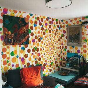 Peut inclure: Un salon avec du papier peint à pois colorés et des œuvres d'art. Un canapé turquoise avec un coussin orange et une couverture à rayures est au premier plan. Un ordinateur portable est posé sur la couverture. Une lampe gris foncé est suspendue au plafond.