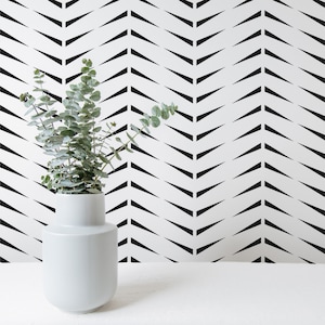 Könnte beinhalten: Eine weiße Keramikvase mit abgerundeter Form, gefüllt mit grünen Eukalyptusblättern, steht vor einer weißen Wand mit einem schwarzen Chevron-Muster.