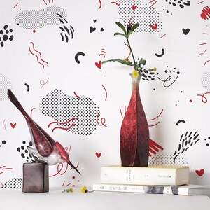 May include: A white wall with a black and red geometric pattern. Two red bird sculptures are on a table in front of the wall. A red vase with a single branch of flowers is on a stack of books.