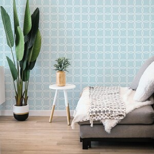 Op de afbeelding: Een lichtblauwe bank met een wit en grijs gestreept deken, een kleine witte bijzettafel met een gouden vaas en een groene plant, en een grote groene plant in een zwart-witte pot. De muur achter de bank is bedekt met een lichtblauwe behang met een wit geometrisch patroon.