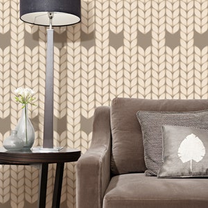 Op de afbeelding: Een bruine bank met twee kussens, waarvan één met een wit bladontwerp, staat voor een muur met een beige gebreid patroonbehang. Een zwarte lamp met een witte kap staat op een ronde houten tafel, naast een vaas met witte bloemen.