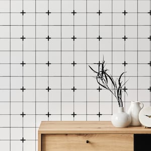 May include: A wooden dresser with a white ceramic vase with dried branches and a white ceramic clock on top. The wall behind the dresser is white with a black grid pattern.