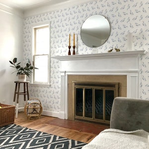 Puede incluir: Una chimenea blanca con una rejilla de metal dorado y una repisa blanca. La repisa tiene un espejo redondo y dos candelabros amarillos. La pared detrás de la chimenea está cubierta con papel tapiz azul y blanco con un patrón de pájaros.