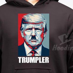 Può includere: Una felpa con cappuccio marrone scuro con un ritratto di un uomo con baffi e capelli stilizzati, su pannelli rossi e blu. La parola "TRUMPLER" è stampata in lettere bianche.