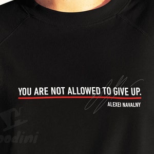Peut inclure: Sweat-shirt noir avec l'inscription blanche "YOU ARE NOT ALLOWED TO GIVE UP." et une ligne rouge en dessous. Le nom "ALEXEI NAVALNY" est en dessous en blanc, avec une signature au-dessus.