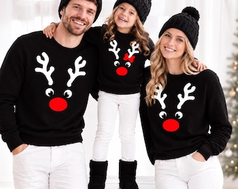 May include: Three black crewneck sweatshirts featuring a white reindeer antler graphic and a bright red nose. Each sweatshirt is paired with white pants and a black knit beanie with a pom-pom. The child's sweatshirt includes a small red bow.