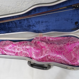 Puede incluir: Un estuche de violín con exterior rígido gris y forro interior de felpa azul. Dentro, un violín está cubierto por una tela rosa brillante con un detallado patrón de cachemira plateado. Un arco de madera está colocado en la parte superior.