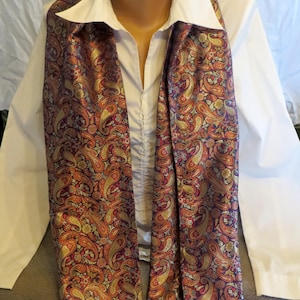 Puede incluir: Una bufanda de cachemira en tonos burdeos, dorados y naranjas, drapeada sobre una camisa blanca con cuello. La bufanda presenta un intrincado patrón de motivos en forma de lágrima. La camisa tiene una parte delantera abotonada.