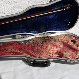 Puede incluir: Un estuche para violín con exterior oscuro y forro interior de terciopelo azul. El estuche contiene un arco de madera asegurado en la tapa y un violín cubierto por una tela con estampado de cachemira en rojo y dorado. Tiene un asa negra.