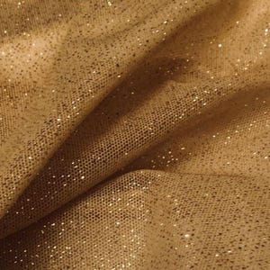 Puede incluir: Primer plano de una tela brillante de color dorado con una fina textura de malla. La tela está drapeada, creando pliegues y sombras suaves. Pequeñas motas brillantes están dispersas por la superficie, agregando un toque de glamour y brillo.