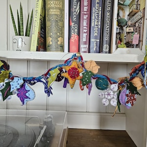 May include: A vibrant, handmade garland featuring star, leaf, and ball shapes in diverse colours. The garland is strung on a twisted cord, suspended before a shelf with books and a small potted plant. A decorative item for the home.