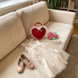 May include: A cream-coloured dress with floral embroidery, ballet flats, a heart-shaped purse, and a floral headband are arranged on a beige sofa. A small tiara and the text "Matching Set" are also visible. The items are from the bloom girl collection.