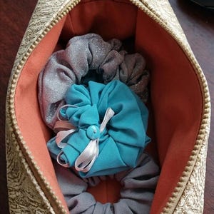 Puede incluir: Un neceser beige estampado con cremallera, abierto para revelar varias gomas para el pelo. Las gomas para el pelo son en tonos grises y verde azulado, con una goma verde azulado atada con una cinta blanca y un botón. El interior de la bolsa es naranja.