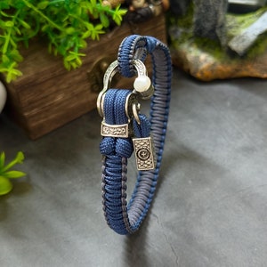 Puede incluir: Un brazalete de paracord azul oscuro con detalles metálicos plateados y una pequeña perla blanca. El brazalete tiene un cierre de broche.