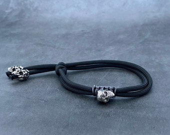 May include: A black paracord bracelet with two silver skull beads. The bracelet is tied with a knot and the beads are positioned at the ends of the bracelet.