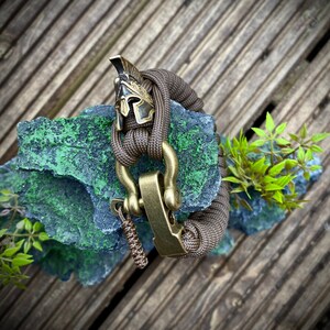 May include: A brown paracord bracelet with a brass clasp and a gold-colored helmet charm. The bracelet is on a green and gray rock.