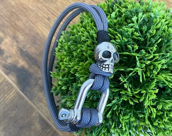 May include: A gray paracord bracelet with a silver skull charm and a silver carabiner clasp.