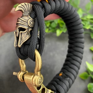 May include: A black paracord bracelet with a gold-toned metal clasp and a decorative Spartan helmet charm.