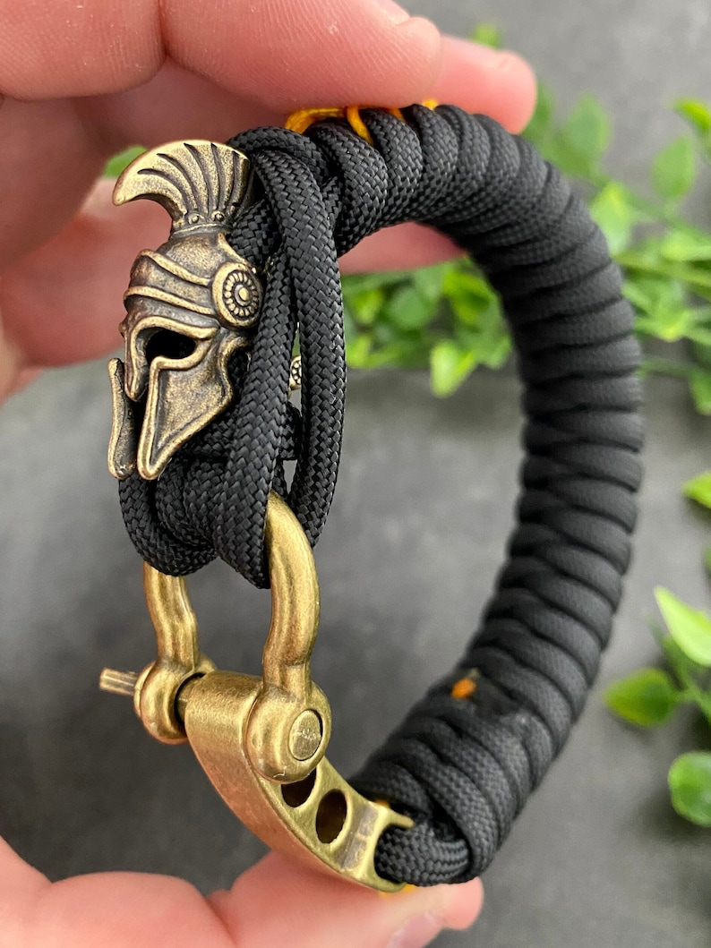 May include: A black paracord bracelet with a gold-toned metal clasp and a decorative Spartan helmet charm.