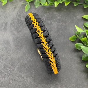May include: Black paracord bracelet with a yellow stitched design. The bracelet is woven with a baseball stitch pattern.