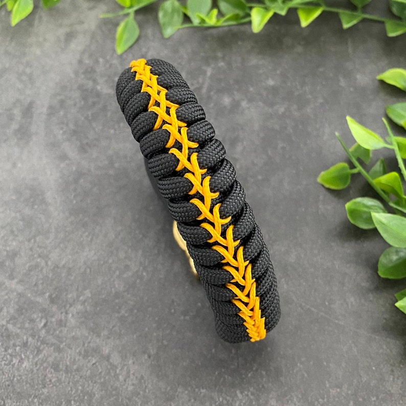 May include: Black paracord bracelet with a yellow stitched design. The bracelet is woven with a baseball stitch pattern.