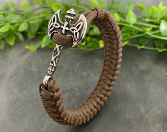 May include: A brown paracord bracelet with a silver axe charm. The axe has a Celtic knot design and a cross on the handle.
