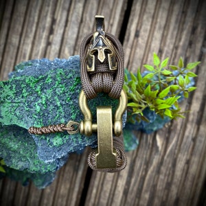 May include: A brown paracord bracelet with a gold metal clasp and a small, black and gold helmet charm.