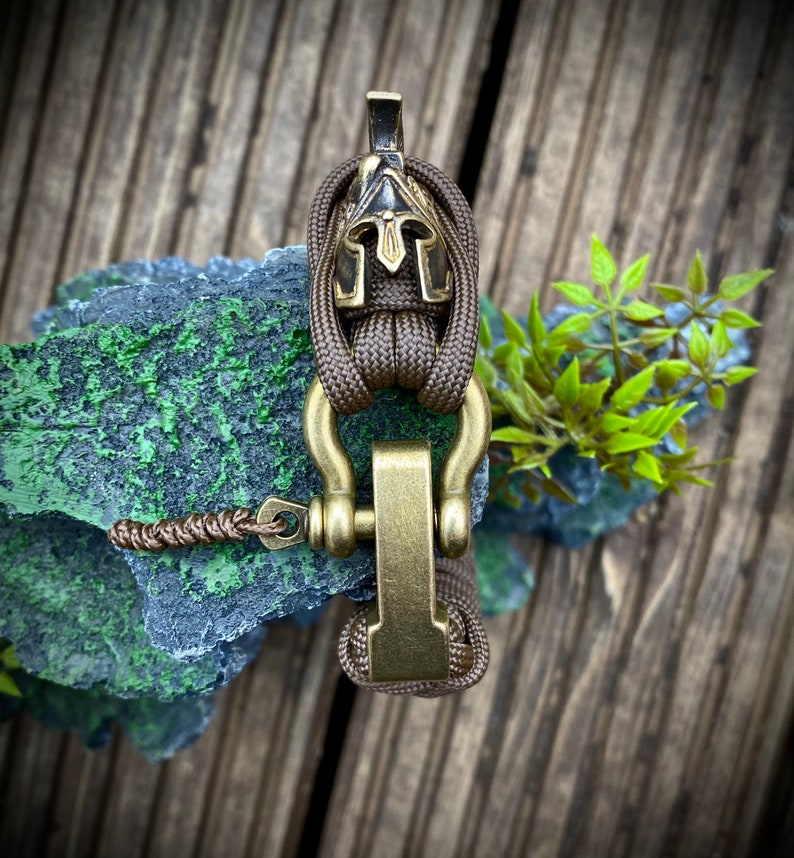 May include: A brown paracord bracelet with a gold metal clasp and a small, black and gold helmet charm.