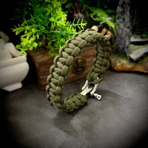 May include: A green paracord bracelet with a silver clasp. The bracelet is made with a cobra stitch pattern.