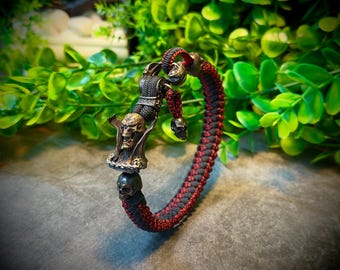 May include: A black and red braided bracelet with a skull and figure charm. The charm features a detailed skull design and a figure with a dark finish. The bracelet is made of braided cord.