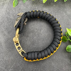 May include: Black paracord bracelet with a gold metal clasp featuring a warrior helmet design. The bracelet has a yellow accent.