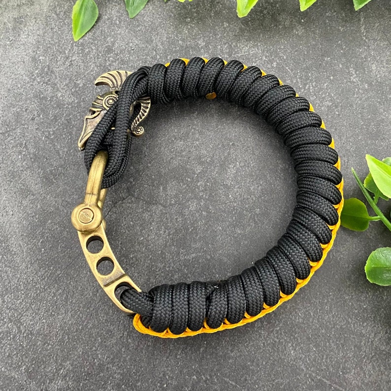 May include: Black paracord bracelet with a gold metal clasp featuring a warrior helmet design. The bracelet has a yellow accent.