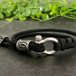 May include: Black paracord bracelet with a silver metal clasp and a silver bead with a symbol on it.