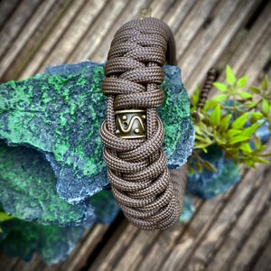 May include: A brown paracord bracelet with a bronze metal bead featuring a geometric design. The bracelet is on a wooden surface.