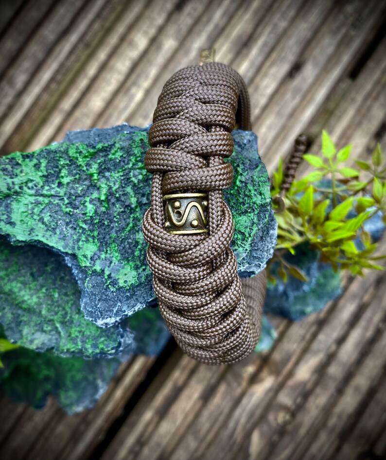 May include: A brown paracord bracelet with a bronze metal bead featuring a geometric design. The bracelet is on a wooden surface.