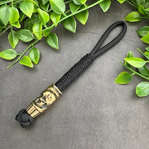Puede incluir: Un llavero de paracord negro con una cuenta de calavera de latón. La calavera está detallada con intrincados tallados.