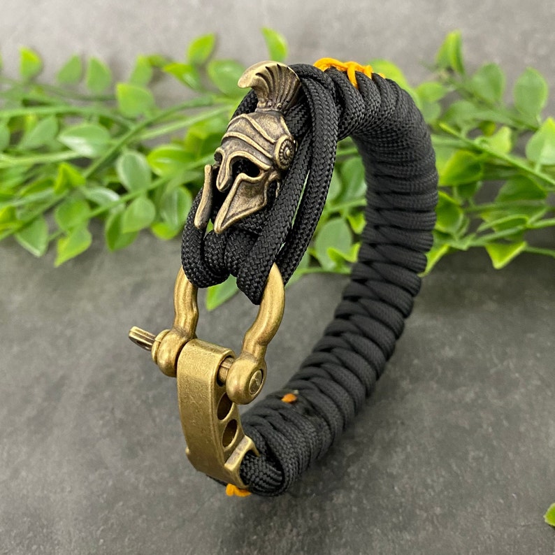 May include: Black paracord bracelet with a gold-toned metal clasp and a Spartan helmet charm.