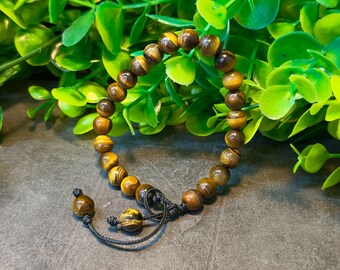 May include: A beaded bracelet made of tiger's eye stones. The bracelet features round, polished beads in shades of brown and gold, strung together on a black cord. The bracelet is displayed against a backdrop of green foliage.