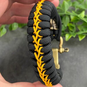 May include: Black paracord bracelet with a yellow stitching detail. The bracelet has a metal clasp.